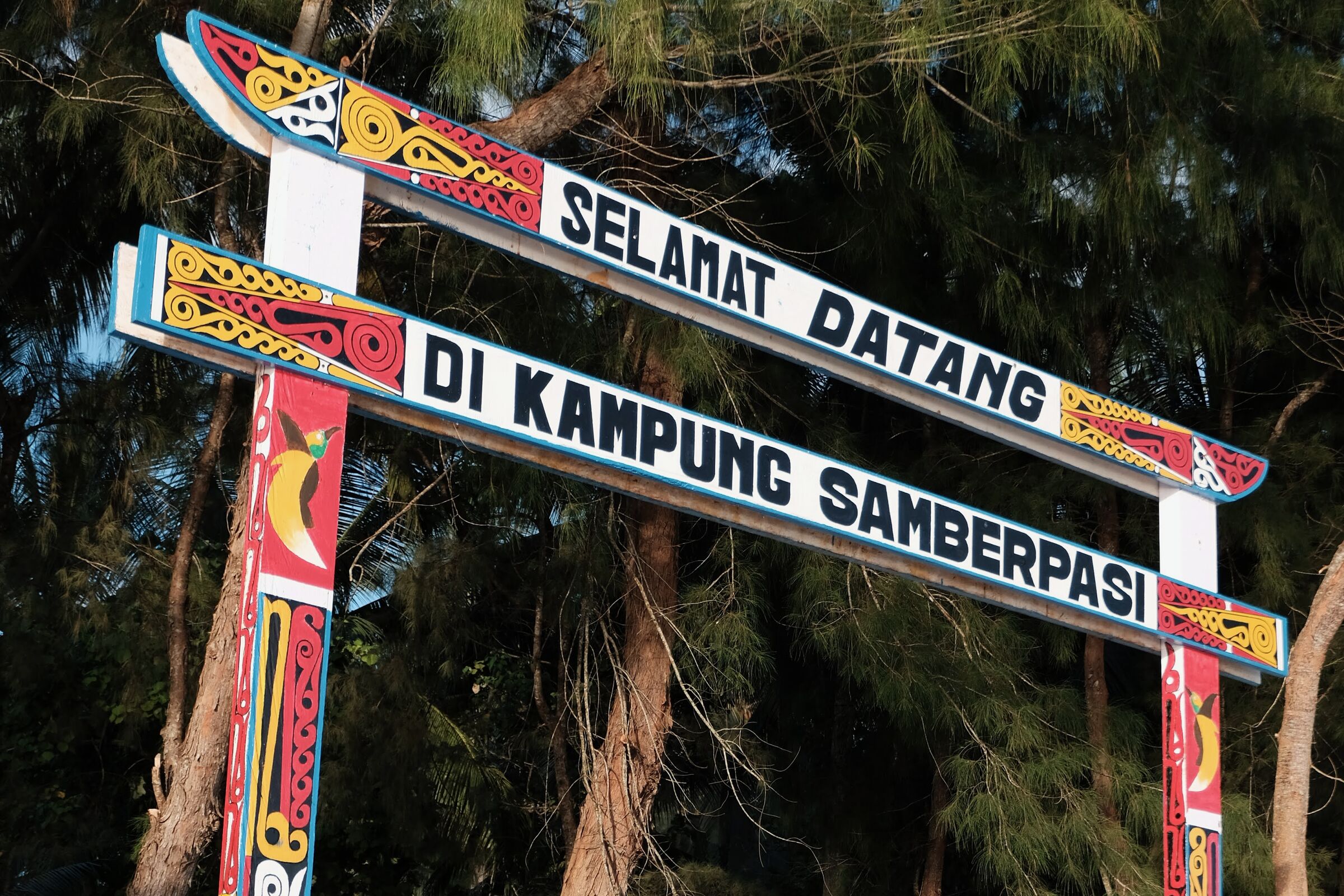 A wooden welcome arch in traditional Papuan colors, reading SELAMAT DATANG DI KAMPUNG SAMBERPASI