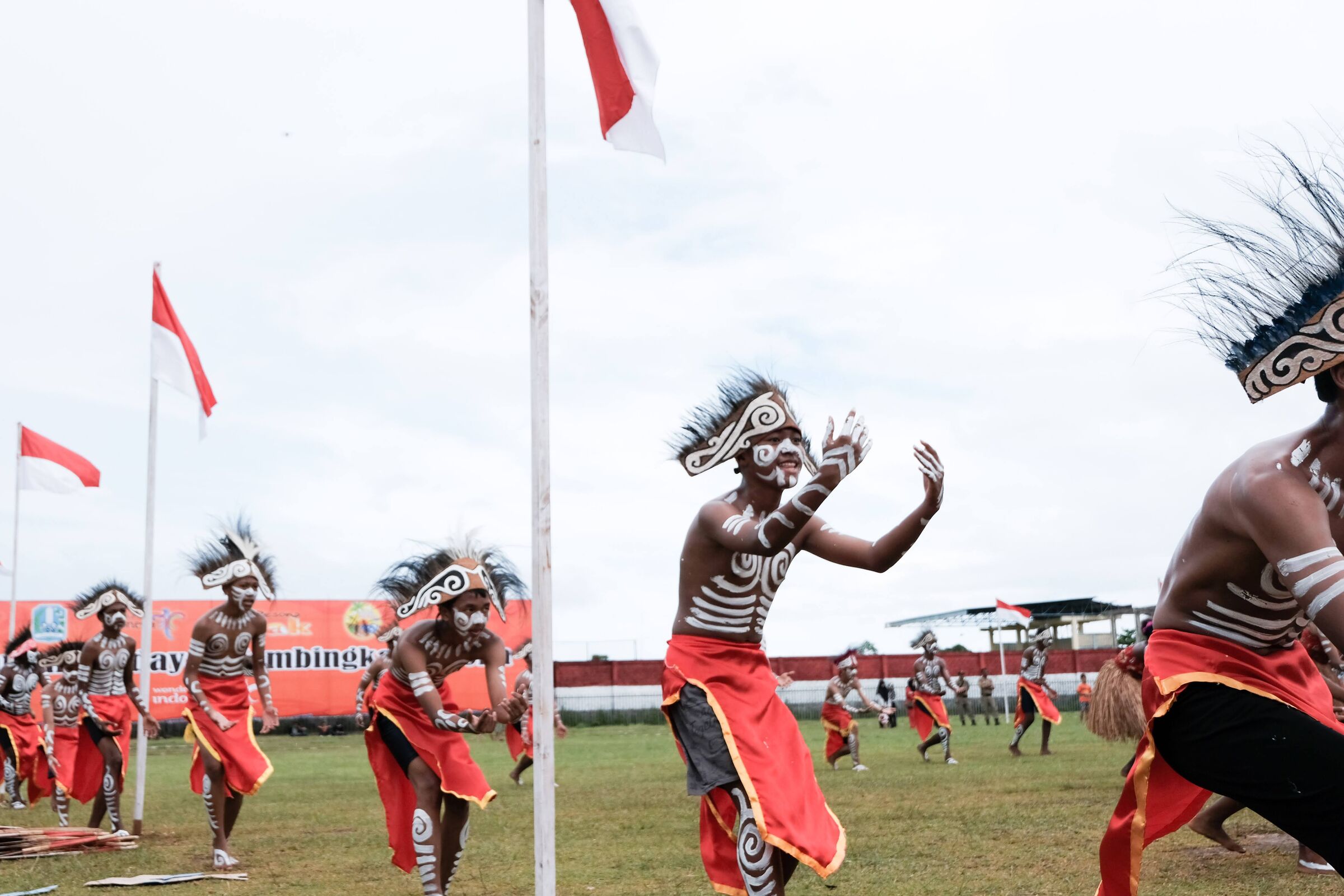 A welcome dance with kids in white body paint and feathered headdresses, mid-formation, with Indonesian red-and-white flags flying behind them