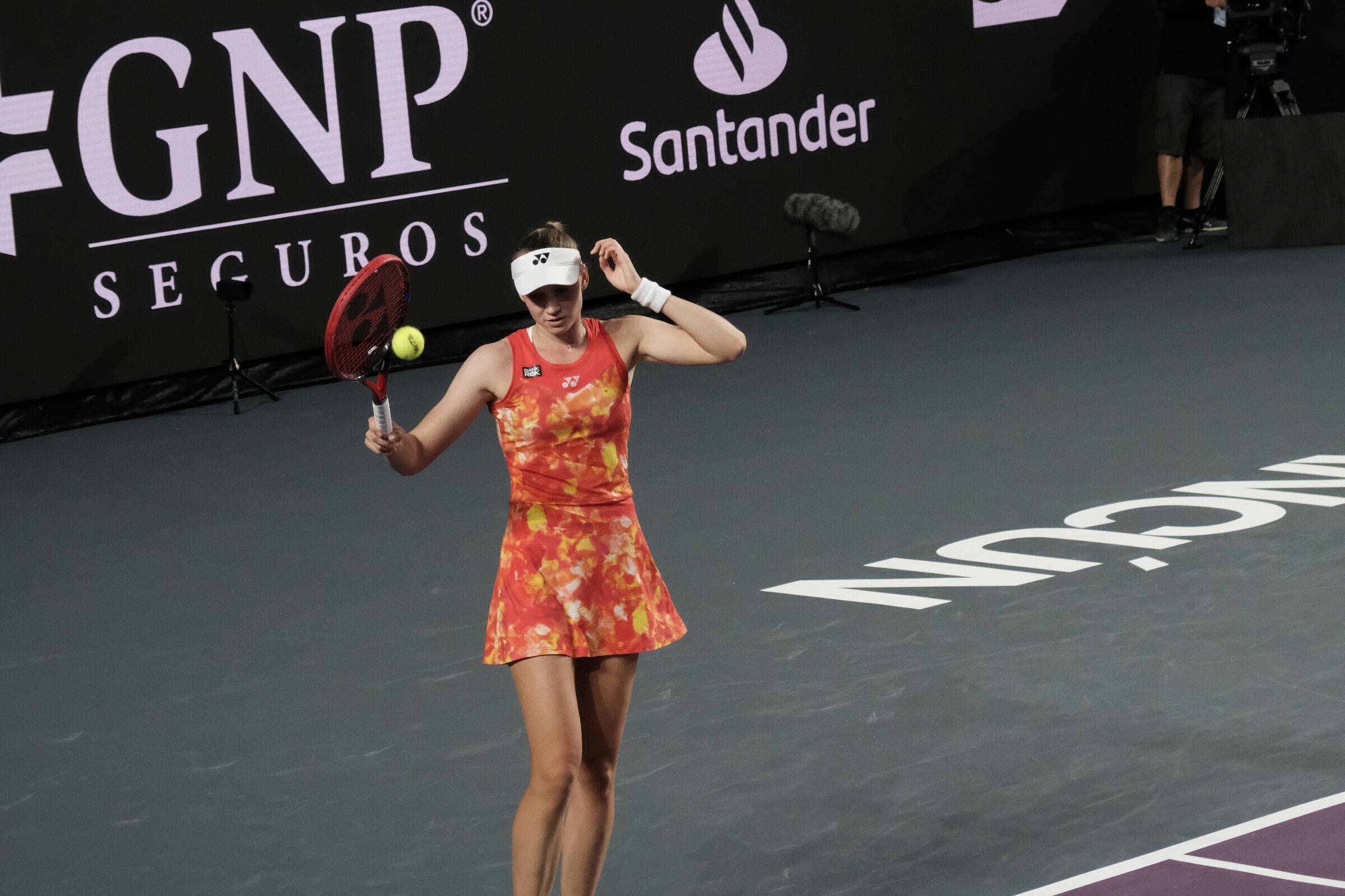 Player in a coral patterned dress and white visor against a dark sponsor wall