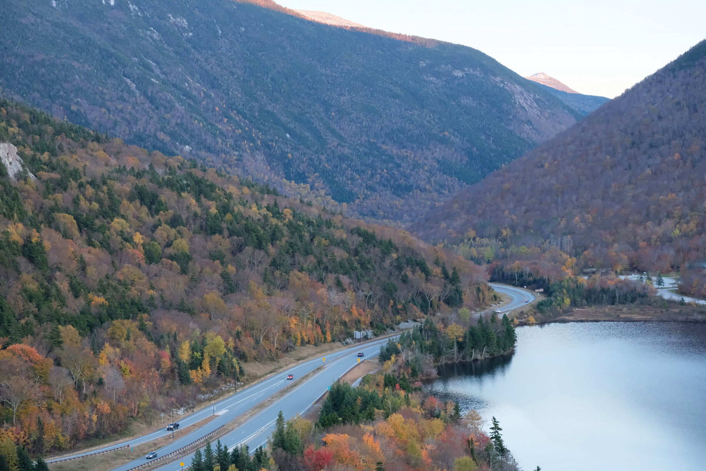 A winding highway through a deep mountain notch, fall foliage on the slopes, a calm lake to the right
