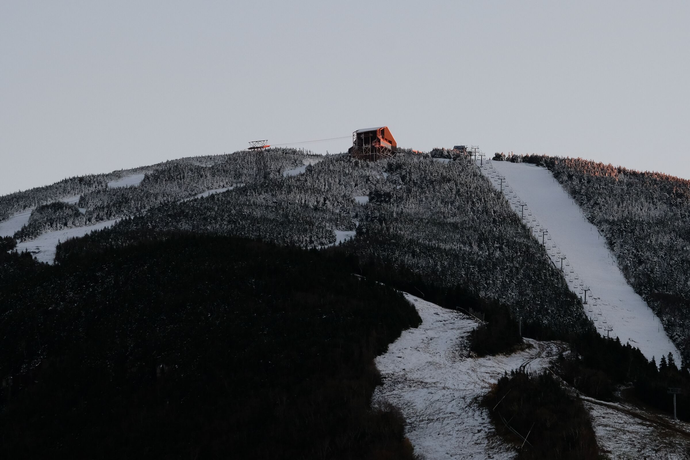 A ski mountain at dusk, snow on the upper trails, a summit lodge silhouetted against a fading sky