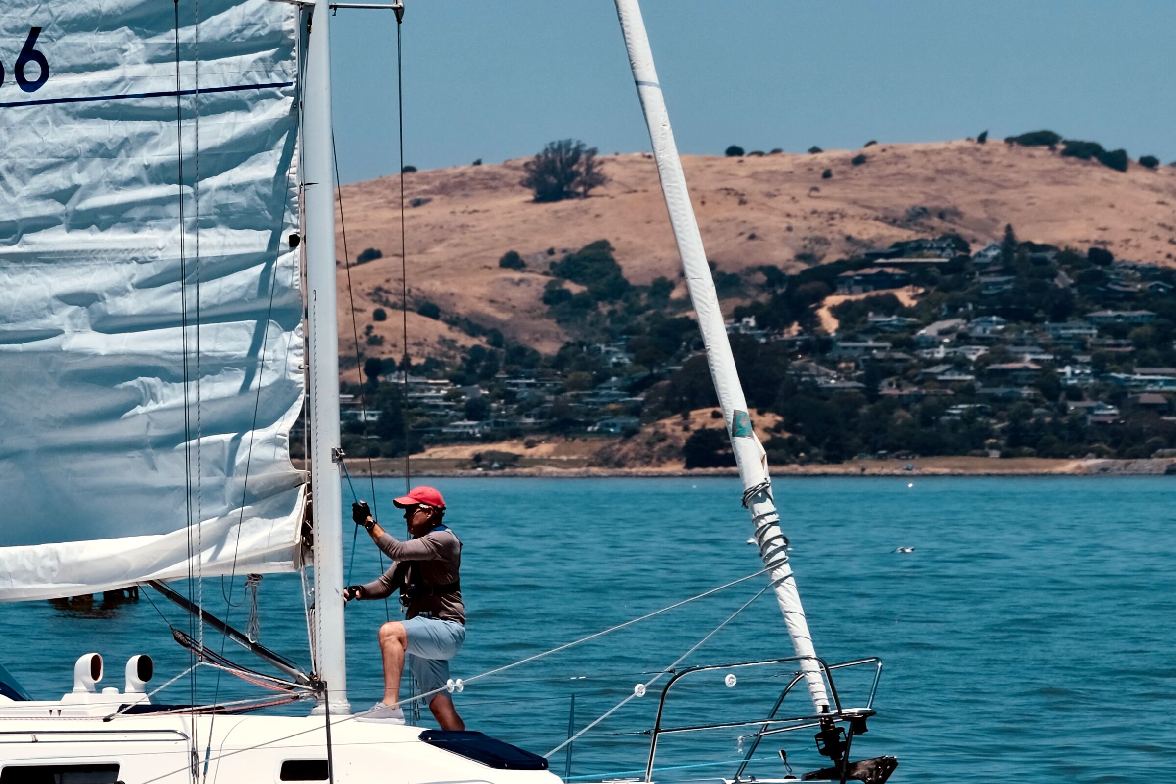 A man in a red cap working the rigging of a sailboat with the bay behind